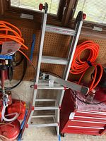 Full view of folded aluminum ladder leaning against pegboard wall near orange extension cords and red toolbox.