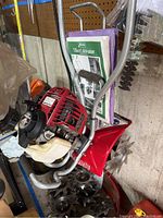 Mantis red and silver gas-powered tiller with manual attached to handlebars showing engine, handlebars, and some tiller blades at bottom.