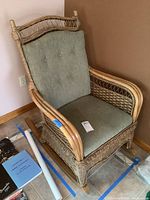 Full view of the rattan and wicker rocker chair with green cushioned seat and back cushions placed next to a wall and tiled floor.