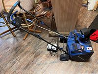 Full view of blue Snow Joe snow blower with two batteries plugged in and charger lying on floor next to it