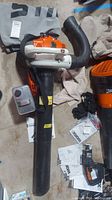 Full view of Stihl SH57C blower with attached long black 44in tube and gray collection bag, showing orange and white motor housing and black carrying strap.
