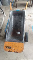 Overall view of the metal wheelbarrow showing the grey metal tub and orange front panel with 'WORX' embossed logo. Two rubber tires visible.