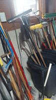 Broom, shovels, rakes, forks, and other garden tools leaning against a wall in garage.