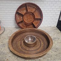 Full view of the two round trays displayed on a kitchen counter. The rattan chip and dip tray is in front with the glass bowl visible, and the wooden divided snack tray is standing upright behind it.