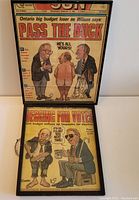 Photo showing both framed newspaper cartoon covers with one frame black metal and the other dark brown wooden, with visible political cartoons and headlines.