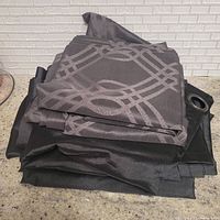 Folded dark grey/brown blackout curtain panels with geometric lattice pattern over black curtains, showing curtain grommet for hanging