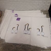 Stack of 6 white cotton placemats each with black, white, and yellow bird designs and fringed edges.