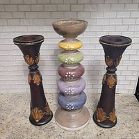 Three candle holders displayed on countertop: one tiered farmhouse pillar holder with multicolored rounded tiers and floral designs, flanked by two dark brown taper holders with gold detailing.