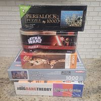 Five boxed games/puzzles stacked: Wrebbit Perfalock 1000-piece puzzle (Twin Towers), Star Wars puzzle, 1000-piece giraffe image puzzle, MonToy 1000-piece cityscape puzzle, The Big Bang Theory Ultimate Genius game.