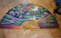 Full view of large hand painted tropical Asian bamboo folding fan wall art laid flat on floor, showing vibrant painted scene of huts, palm trees, and water with boats.