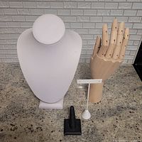 Front view of white fabric necklace display with wooden articulated hand, black ring stand and white earring display on granite countertop in front of subway tile backsplash.