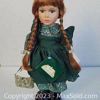 Front view of the porcelain doll showing red braided hair, green floral dress, and apron with original tag attached.