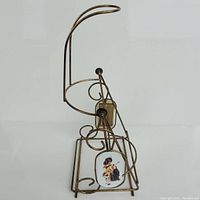 Front view of vintage swinging bottle holder cradle displaying metal wire construction and decorative plaque with image.