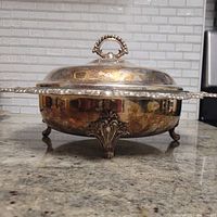 Side view showing ornate feet and overall shape of the warming serving dish with lid and finial handle.