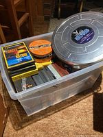 Plastic bin filled with various CDs and DVDs, metal film reel container on top, showing the assortment of media.