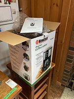 Newfast digital coffee maker in original box with instruction manual on top, seen inside a cardboard box on wooden surface.