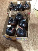 Nine amber glass wine jugs stored in two cardboard boxes on carpet. Jugs have round bodies with handles and black stoppers.