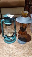 Photo showing side-by-side green metal camping lantern (DIETZ) and amber glass oil lantern with metal base and top.