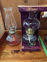 Two oil lamps; one with ribbed red glass base and clear chimney, second in original box labeled Romance Collection with rounded glass base.