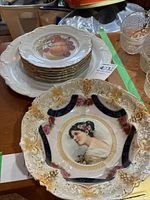 Stack of six plates with fruit design and gold trim, with large decorative platter featuring a woman's portrait and ornate gold and blue details beneath.