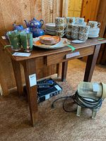 Wooden table with drawer in worn condition. Table holds various ceramic and glassware items but contents not included. Heavy and sturdy with a rustic wooden finish. Shown from side and front angle with drawer partially open.