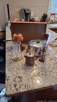 All items shown on granite countertop: copper fondue set with six forks on stand, metal coffee pot, glass candle holder with brass base, silver cake stand with dome lid.