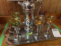 Full view of metal decanter on tray with five colored glass stemmed glasses in green, amber, and orange hues, on a decorative metal tray with handles.