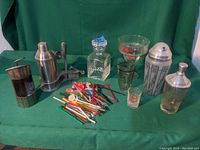 Full lot photo showing glass decanter, metal wine dispenser, metal cocktail shaker, clear shot glass, corkscrew, jigger, and a large collection of colorful vintage plastic stir sticks on green fabric background.