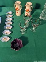 Photo showing two tall cream-colored vases with orange flowers, six small oval blue dishes with floral motifs, three vintage glasses with floral designs, and a small purple flower-shaped dish.