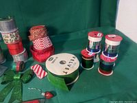 Photo showing multiple rolls of Christmas-themed ribbons including green velvet, red, and white patterned ribbons, along with a red metallic candy cane and green glitter bow on a green background.