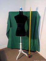 Photo of black velvet covered dress form on white wooden tripod base with measuring tape for size reference and green background.