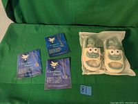 One photo showing three Christmas Peace necklaces with dove and heart pendants lying flat, and one pair of frog-themed slippers in plastic packaging beside the necklaces on a green background.