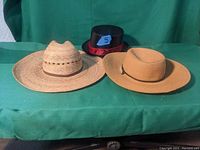 Photo of three hats displayed on green cloth backdrop: two straw hats and one black top hat with red decoration and blue label.