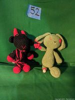 Front view of two knitted dolls on green background, showing black and red devil doll and light green rabbit doll