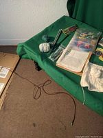 Yarn balls, knitting needles, white knitted pieces, printed patterns, and magazines arranged on green fabric.