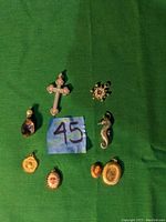 Seven assorted pendants laid out on green fabric, showing rhinestone cross, zodiac, purple glass, porcelain girl locket, and gold-tone pieces.