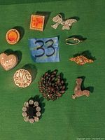 Top-down view of 11 assorted decorative pins arranged on green fabric with blue note labeled '33'. Pins include a heart-shaped mother-of-pearl pin, silver bow pin, multiple ornamental and stone-adorned pins in various shapes and colors.