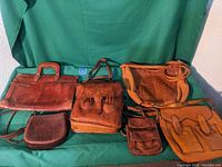 Six vintage leather bags arranged on a green fabric background, showing various sizes and styles.
