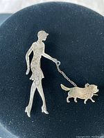 Photo showing entire brooch with lady holding leash attached to dog figurine, laid on black background for contrast.