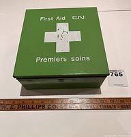 Top view photo of green metal first aid kit box showing white cross and text on lid, alongside ruler for scale.