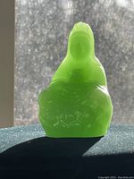 Side/back translucent view of the jade Buddha figurine showing shape and texture details against a textured curtain background.
