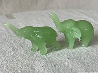 Front and side view of two small green jadeite elephant figurines with raised trunks, placed on a light fabric surface.
