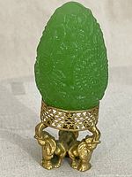 Front view of green jadeite jade egg on detailed solid brass elephant stand with carved floral pattern on egg.
