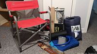 Photo shows Coleman red and grey folding chair, rolled blue sleeping bag, machete with brown leather sheath, black folded item, and boxed items stacked behind.