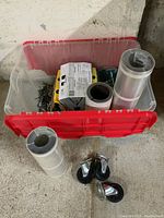 Clear plastic tote with red lid containing metal brackets, chains, flashing rolls, and casters placed in front on floor.