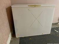 White Kenmore chest freezer against a wall on dark carpeting. Large 'X' mark on front, handle on top with lock, some scuffs and dirt visible.