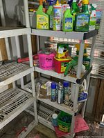 Four-shelf white plastic shelving unit holding multiple bottles, buckets, and jugs of garden chemical products including fertilizers, weed control, pesticides, and herbicides.