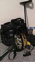 Photo showing two large OGIO black backpacks, green camo pouch, black folding stool, kneeling pad, and guidebook box on floor with walking stick.