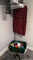Green laundry hamper filled with various laundry and cleaning products, gray bathmat on floor, black shoe forms, burgundy garment bag, and multiple hangers on wall-mounted rod.