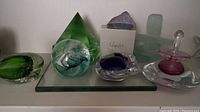 Photo of seven glass paperweights on glass shelf showing various shapes including pyramid, inuksuk, cat, and signed perfume bottle